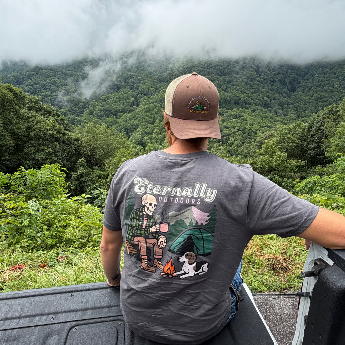A man wearing a brown trucker hat and a gray graphic t-shirt sits in the bed of a pickup truck, looking out at a misty green mountain landscape. The back of the t-shirt features a detailed camping graphic with the brand name 'Biornally Outdoors'.