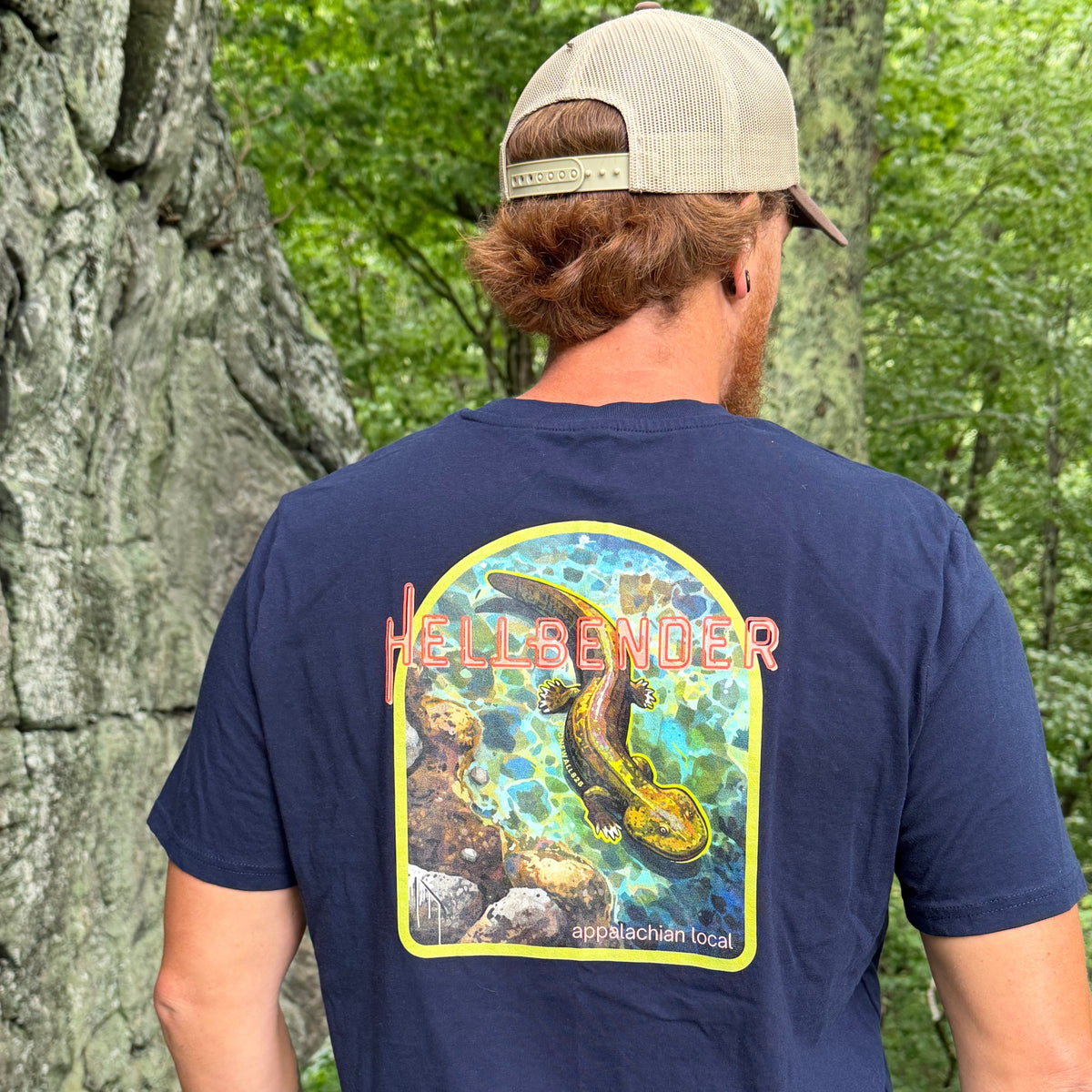 A man wearing a navy blue t-shirt with a large graphic print on the back, photographed from behind in an outdoor forest setting. The graphic features a yellow border and an illustration of a fish with the text 'HELLO ANGLER' in pink.