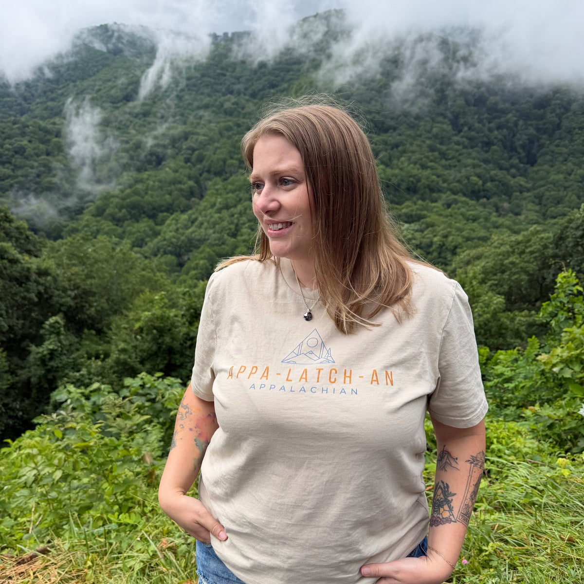 A woman stands outdoors in a lush green mountain landscape with mist, wearing a beige graphic T-shirt that reads 'APPA LATCH-AN APPALACHIAN' and denim shorts.