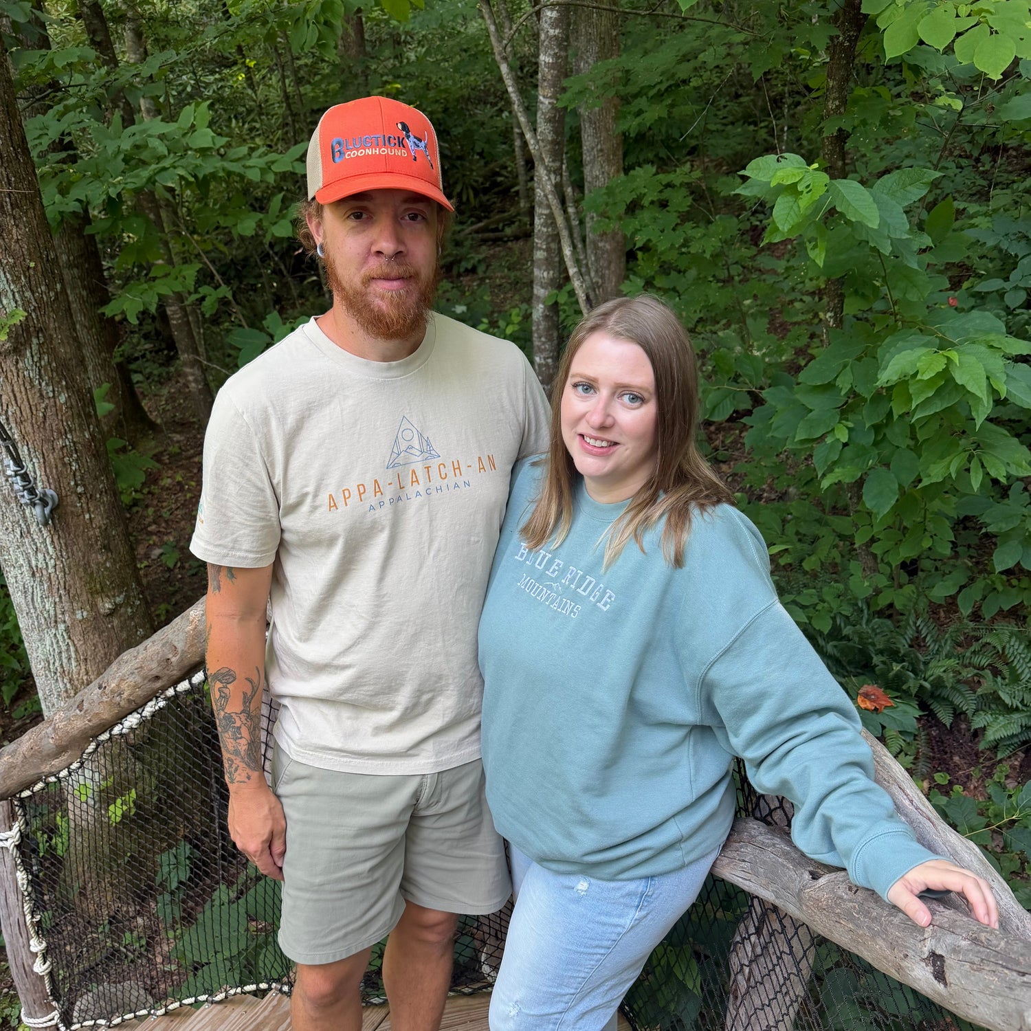 A man and a woman standing together on a wooden boardwalk surrounded by a dense green forest. The man wears a beige t-shirt with 'Upper Catchers' printed on it, a bright orange cap, and sandals.
