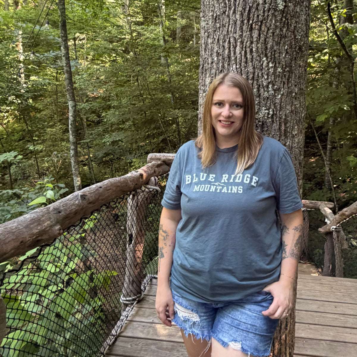 A woman stands on a wooden suspension bridge in a forest, wearing a slate blue t-shirt with 'BLUE RIDGE MOUNTAINS' printed on the front and denim shorts.