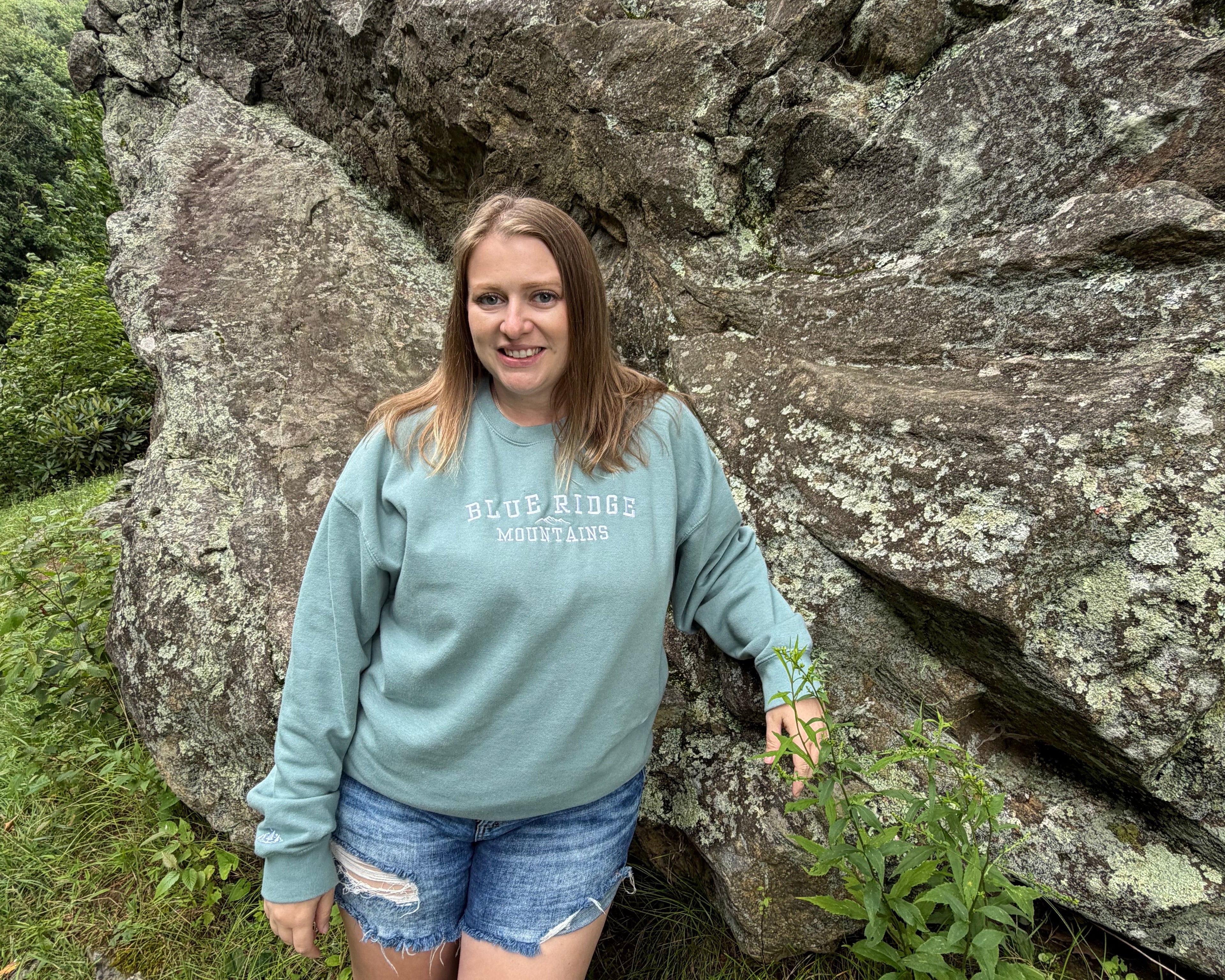 A woman stands outdoors in front of a large rock formation, wearing a sage green sweatshirt with 'BLUE RIDGE Mountains' printed across the chest and denim shorts.