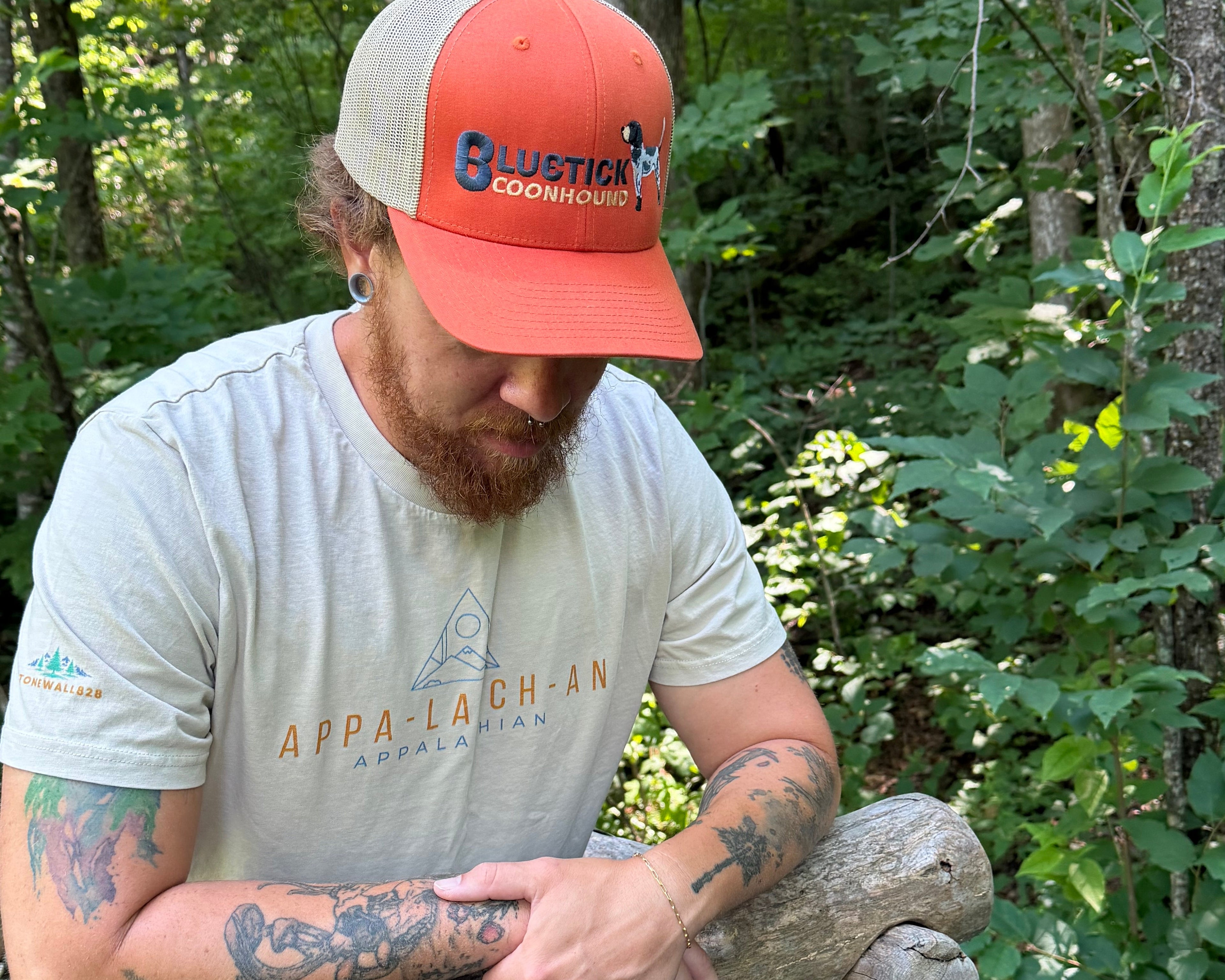 A man in a wooded outdoor setting wearing a beige and orange cap with 'Blustic Construction' printed on it, a light gray t-shirt with 'Appalachian Appalachia' printed on the front, and leaning on a rustic wooden structure.
