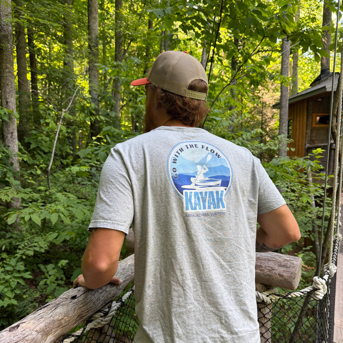 A man wearing a light gray t-shirt with a blue and white 'KAYAK' back graphic, standing on a wooden suspension bridge in a forest.