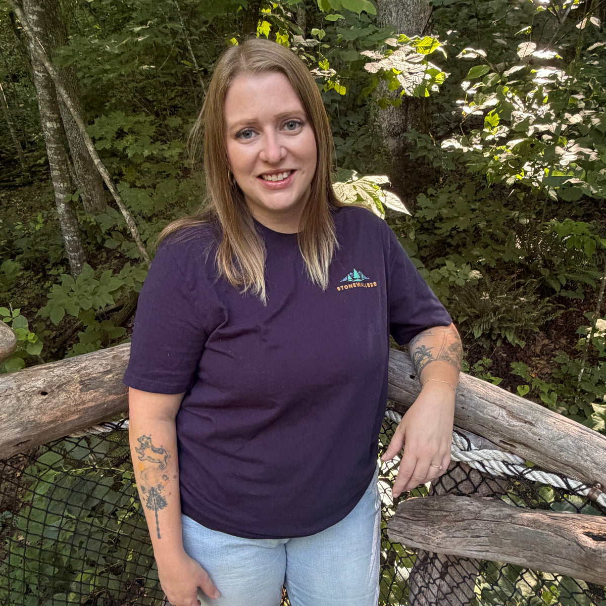 A woman with blonde hair stands on a wooden bridge in a forest, wearing a dark purple t-shirt with a small green and blue mountain logo that reads 'Mountain View' on the left chest, paired with light blue jeans.