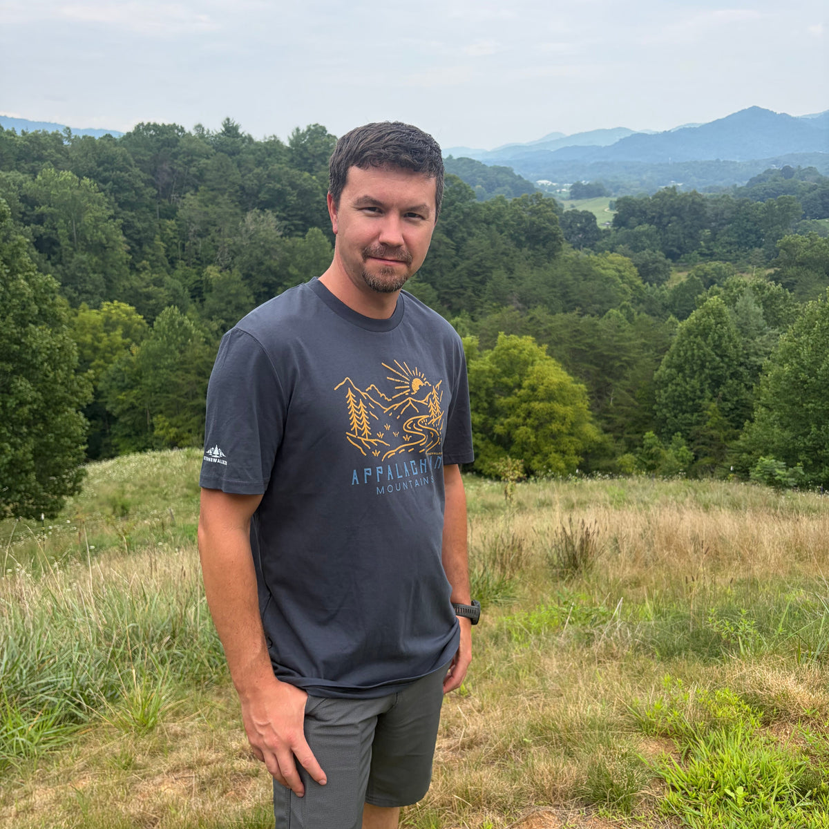 A man standing outdoors on a grassy hill with a forest and mountains in the background, wearing a dark gray t-shirt with a gold graphic and gray shorts.