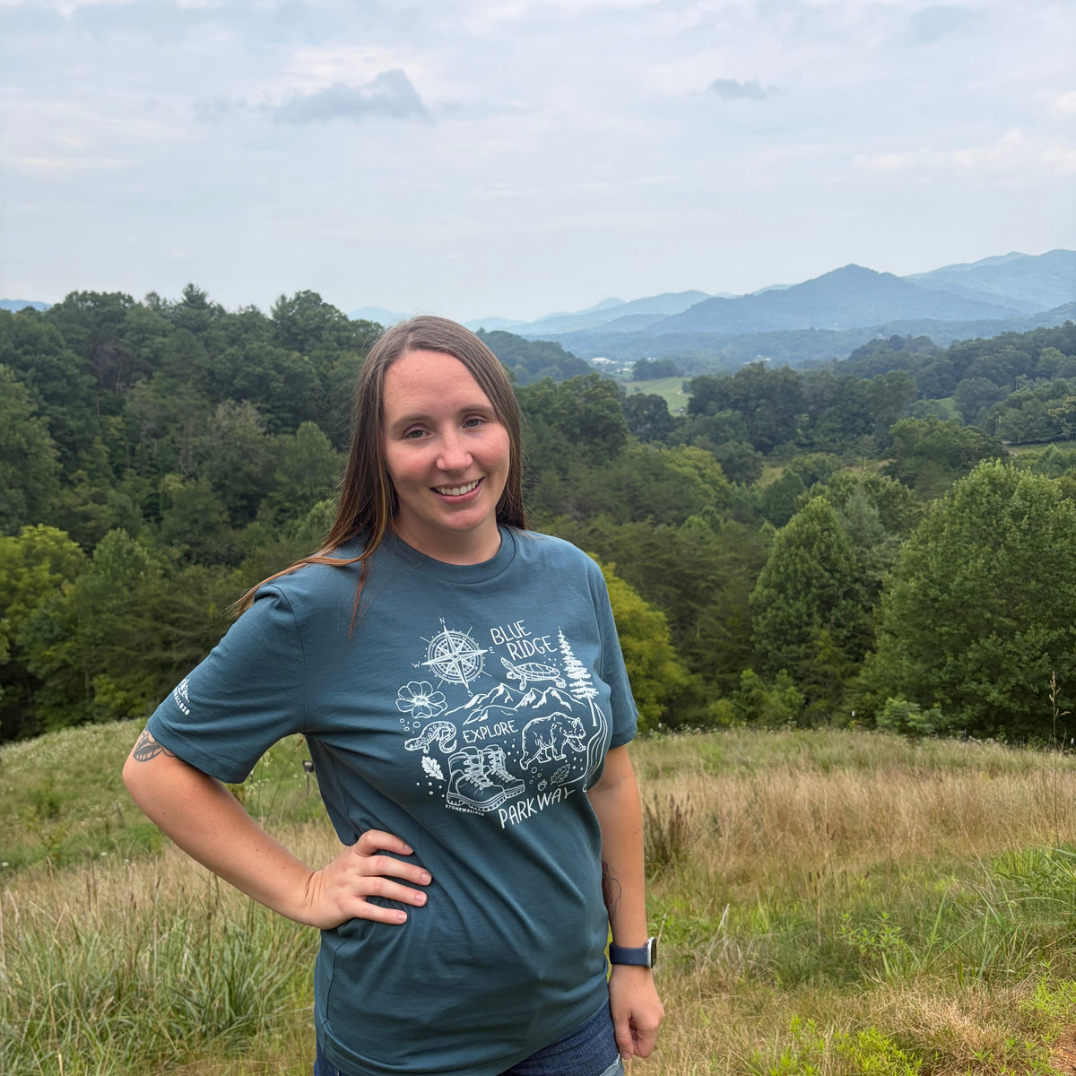 A woman standing outdoors on a grassy hill with a forest and mountains in the background, wearing a dark teal graphic t-shirt and denim shorts.