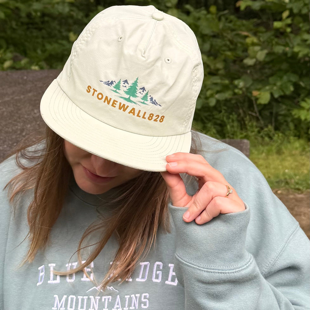 A person outdoors wearing a light beige baseball cap with 'STONEWALL826' and pine tree embroidery on the front, paired with a light blue sweatshirt that reads 'BLUE RIDGE MOUNTAINS'.