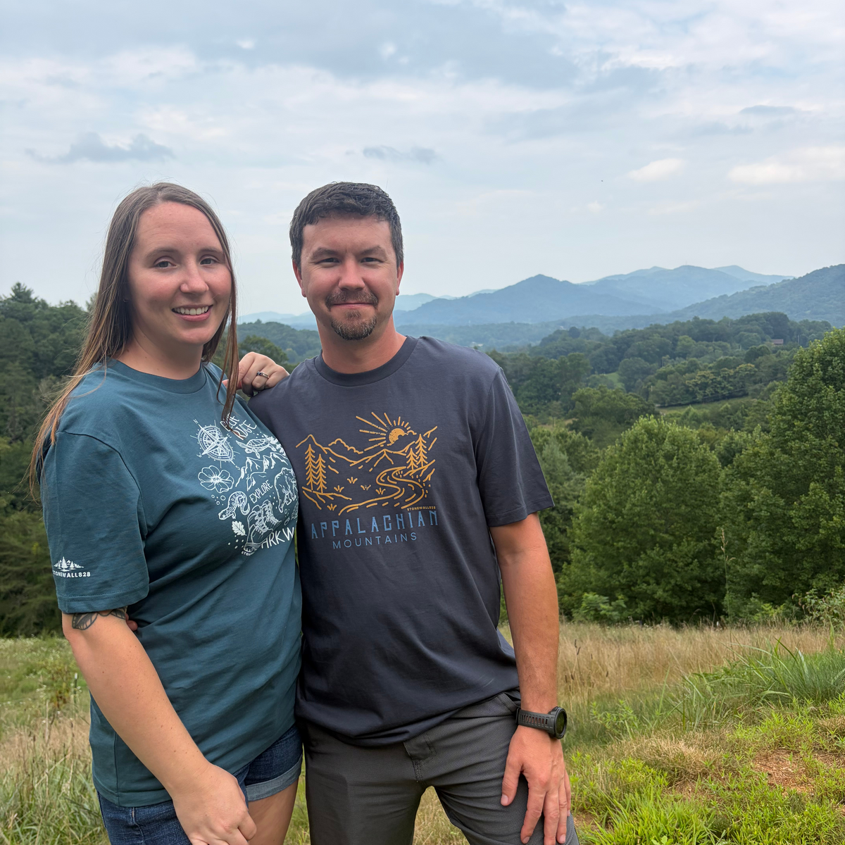 A man and a woman standing outdoors on a grassy hill with mountains and trees in the background. The man wears a dark gray t-shirt with a gold graphic and the words 'Peach Labiche Mountain' printed on the front, paired with gray shorts.