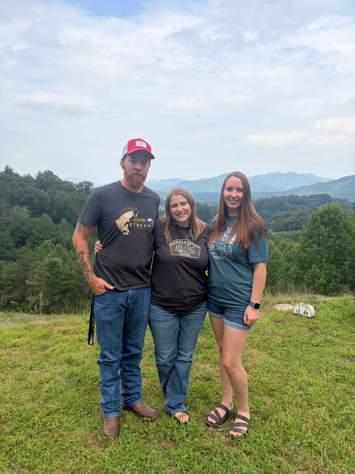 Three people standing together outdoors on a grassy hill with mountains and trees in the background. The man wears a red cap, a dark graphic T-shirt, and jeans.
